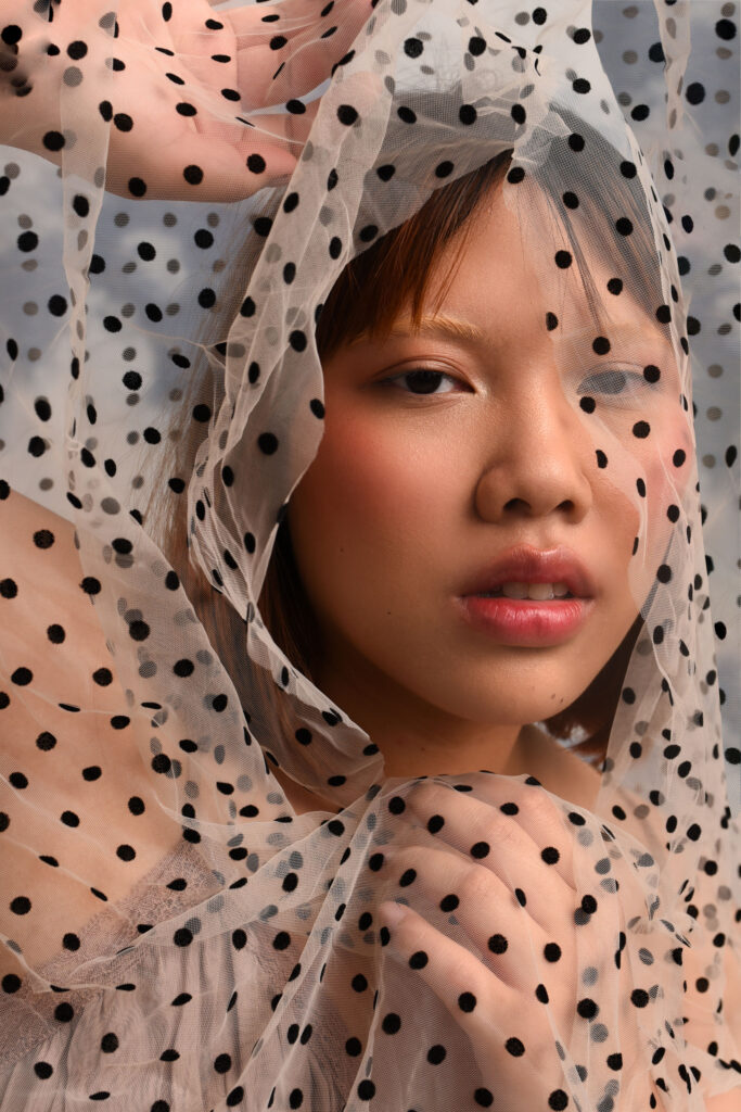 A person with a bleached brow and subtle blush posing with a semi-sheer black and white polka dot fabric framing their face.