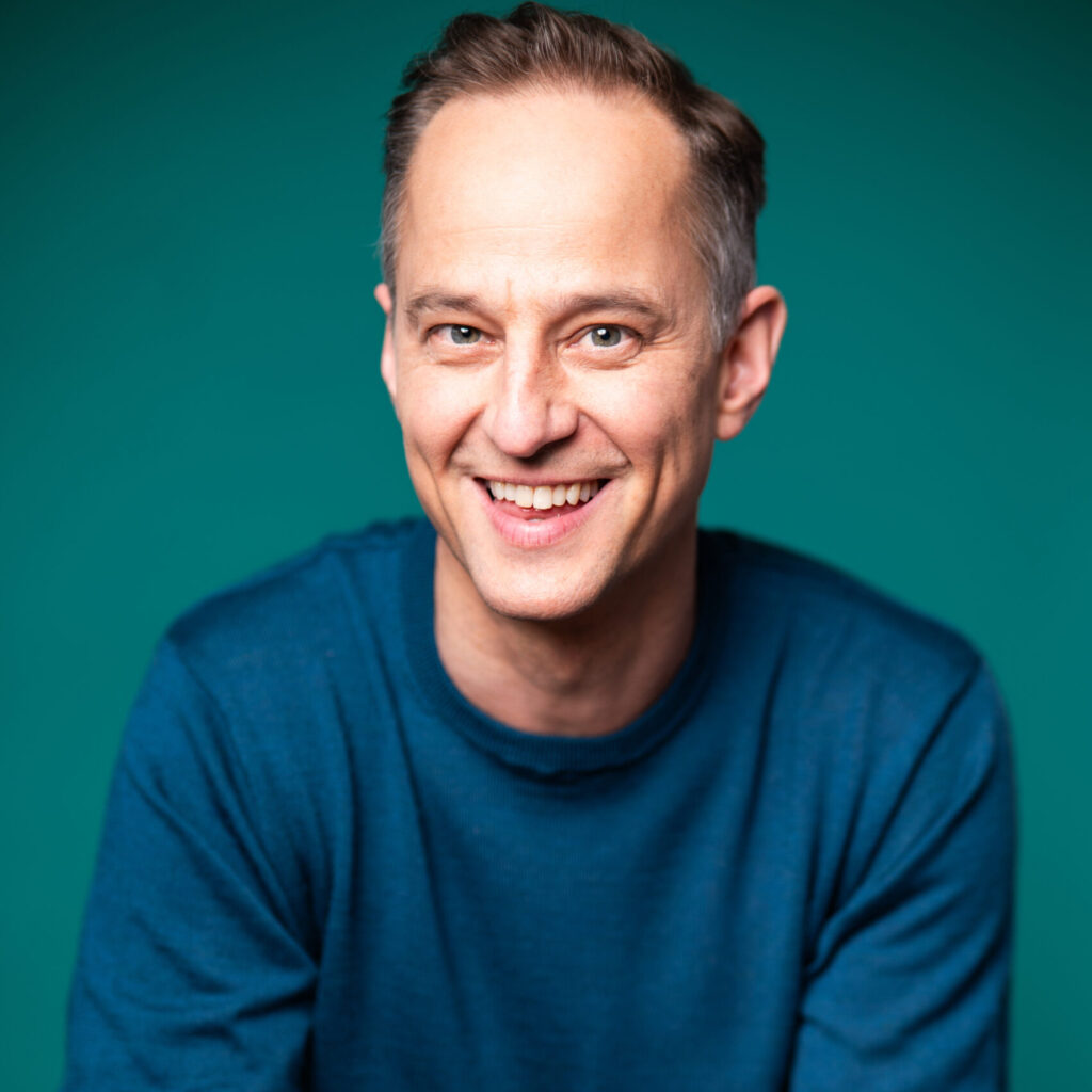 A smiling slim man, with brown hair in a blue sweater.