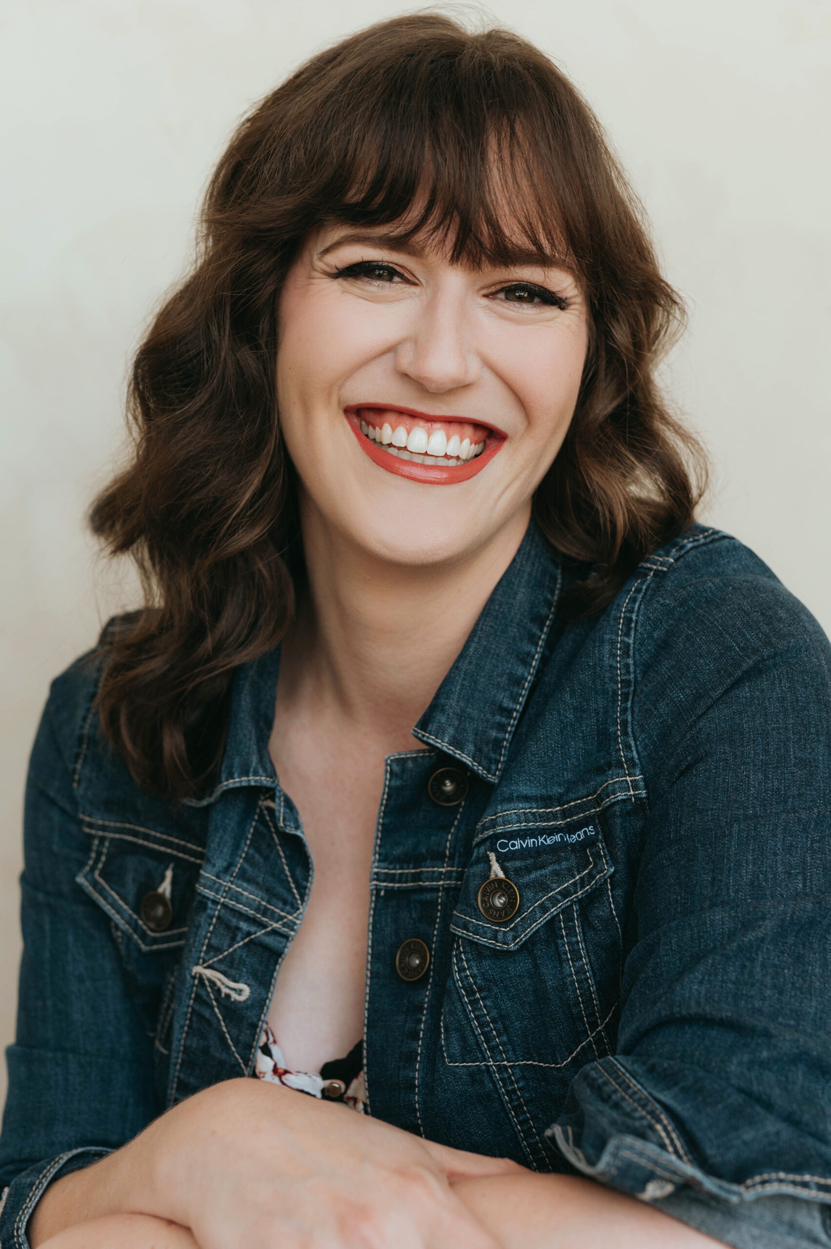 A woman in her 30s with long wavy brown hair smiling brightly. She is dressed in a denim jacket.
