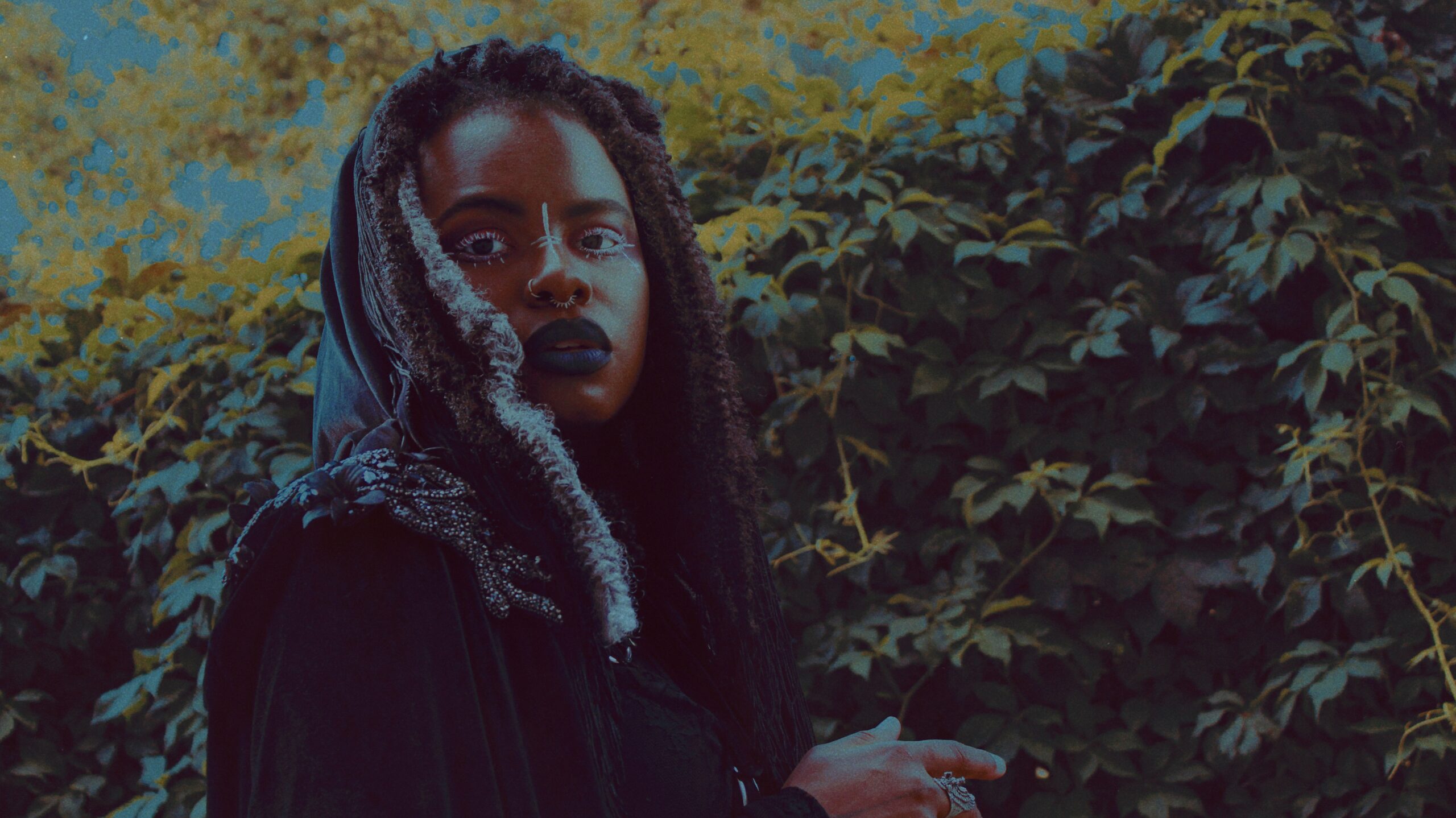 Black woman dressed in a dark cloak and hood with shoulder length black and silver locs looking intently at the camera.