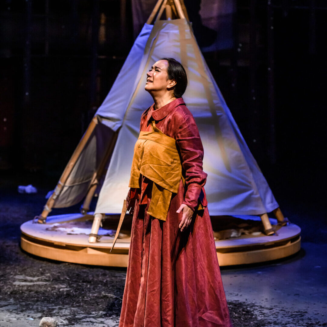 Shanawdithit: A woman dressed in red on a stage, looking upwards with a questioning expression