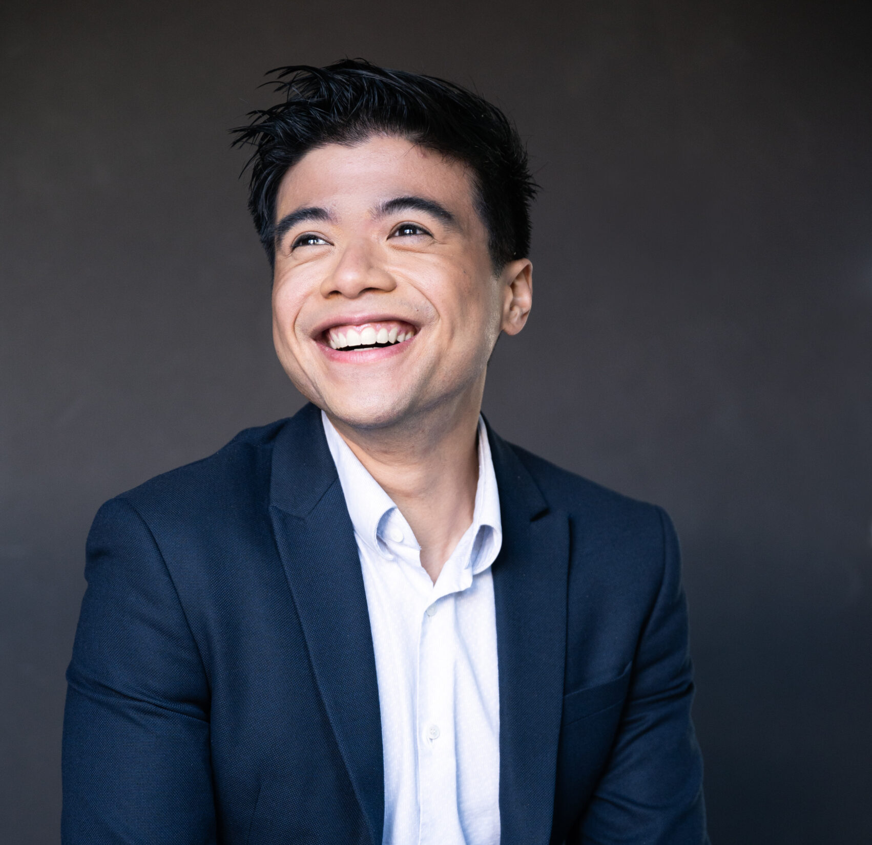 Headshot of Mishael Eusebio, A smiling young man in his 20's with black hair wearing a blue suit, white shirt with no tie, a chain bracelet with pearls in his right hand and a watch on his left.