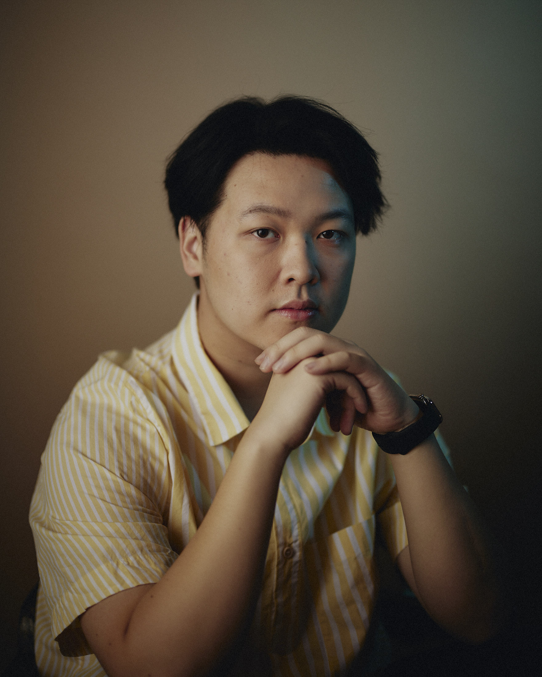 East Asian man with black hair mostly parted to the left side. He's in a striped yellow shirt crouching forward on his hands which are clenched together propped on his knees.