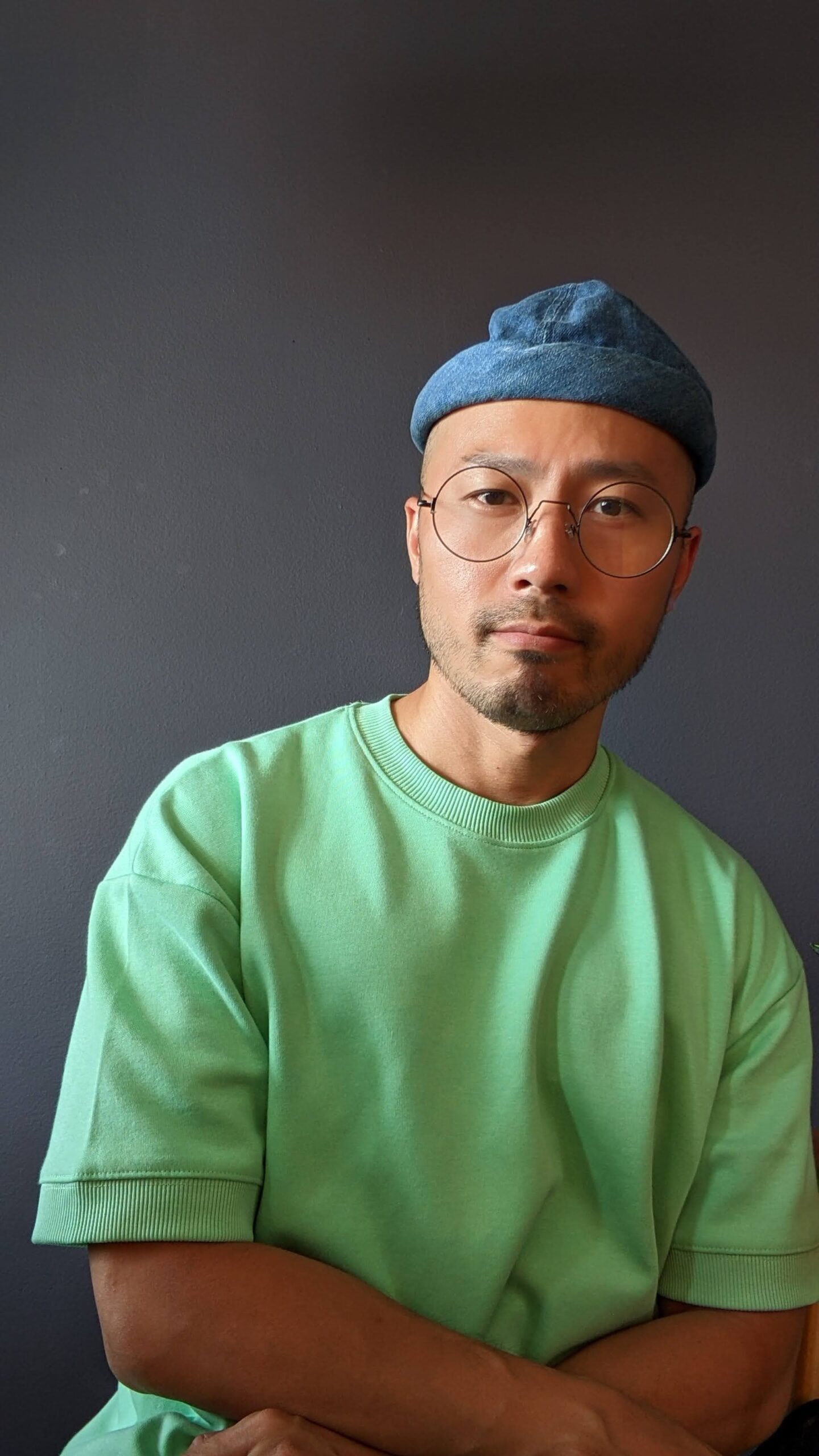 Keith is wearing a blue docker hat, circular glasses and a green sweater