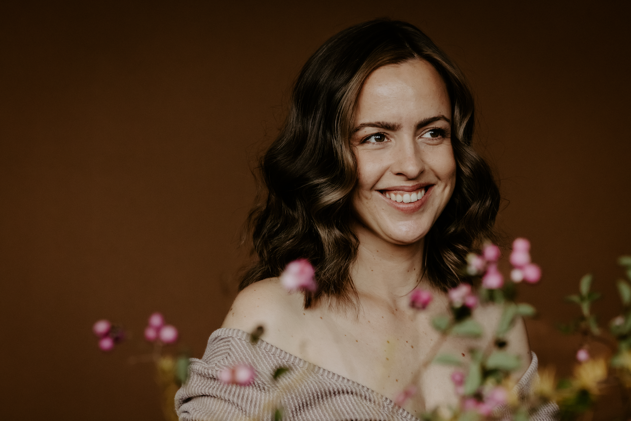 Smiling face of Marie Andrée Gaude with long brown, wavy hair, wearing a beige outfit. There are pink flowers in soft focus in the foreground