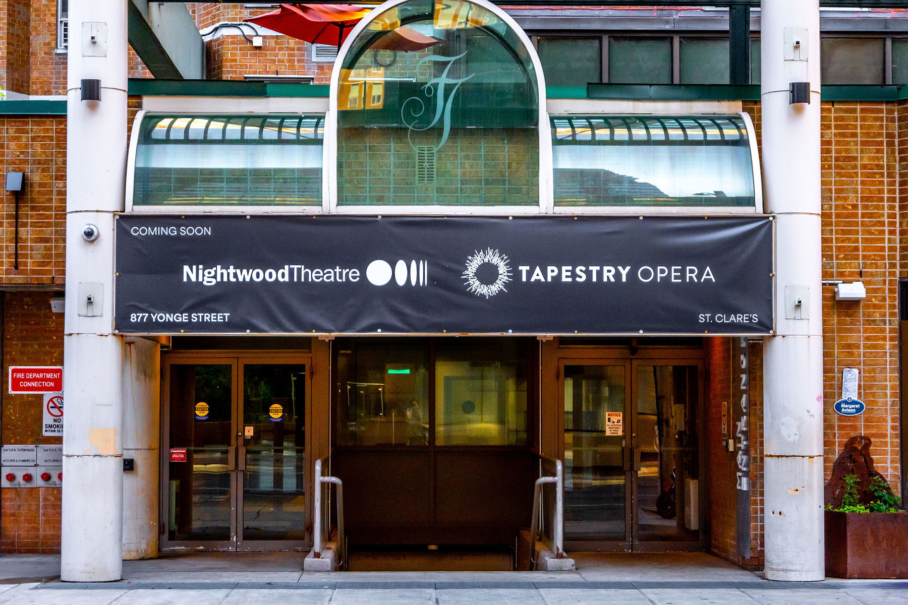 Colour photo of the entrance to a building. A black banner that reads Tapestry Opera and Nightwaood Theatre hangs over the doorway. There is a white column on either side of the entrance. Photo by Dahlia Katz